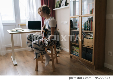 Remote view of talented curly preteen girl sitting on chair and playing on electric guitar, learning new song at home. Pretty kid teaching to play guitar in cozy bedroom with modern interior. Remote view of talented curly preteen girl sitting on chair and playing on electric guitar, learning new song at home. Pretty kid teaching to play guitar in cozy bedroom with modern interior. 95222873