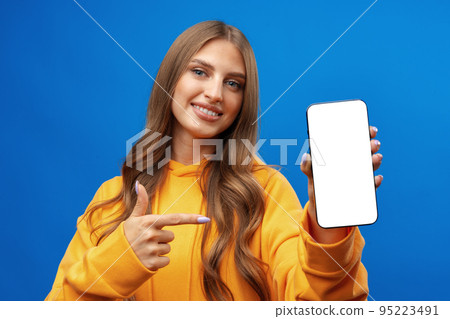 Young woman showing big blank cellphone screen on blue background in studio Young woman showing big blank cellphone screen on blue background in studio 95223491