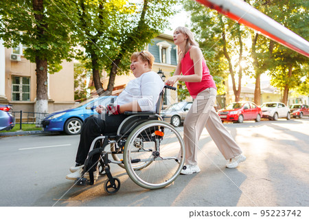 Young female caregiver pushing wheelchair with female person with disability across city street Young female caregiver pushing wheelchair with female person with disability across city street 95223742