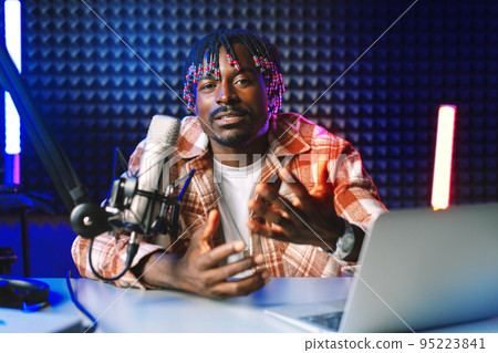 African radio host sitting at desk recording in studio with microphone and laptop African radio host sitting at desk recording in studio with microphone and laptop 95223841