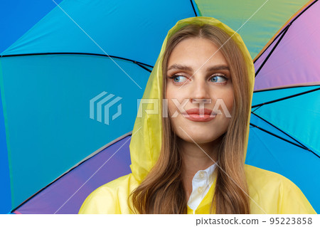 Young woman in yellow raincoat with rainbow umbrella against blue background in studio 95223858