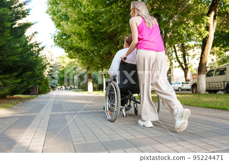Young female caregiver pushing wheelchair with female person with disability across city street Young female caregiver pushing wheelchair with female person with disability across city street 95224471