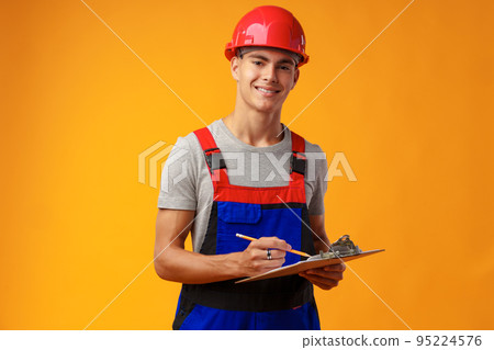 Young construction worker wearing hardhat and holding a clipboard on yellow background in studio Young construction worker wearing hardhat and holding a clipboard on yellow background in studio 95224576