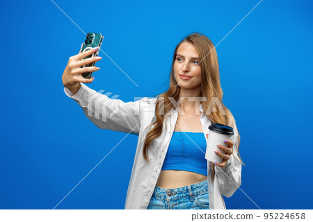 Portrait of smiling beautiful woman with takeout coffee using smartphone against blue background in studio Portrait of smiling beautiful woman with takeout coffee using smartphone against blue background in studio 95224658