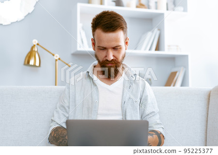 Young bearded man sitting on sofa at home using laptop computer 95225377