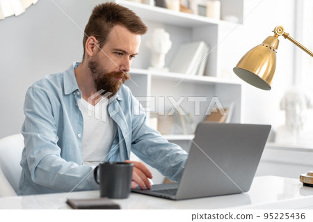 Stylish bearded man working on laptop from home sitting at the table Stylish bearded man working on laptop from home sitting at the table 95225436