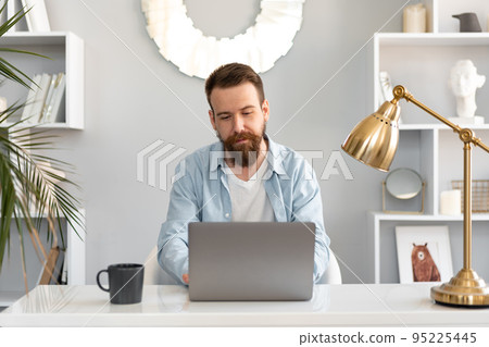 Stylish bearded man working on laptop from home sitting at the table Stylish bearded man working on laptop from home sitting at the table 95225445