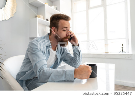 Young bearded businessman working from home sitting at the desk and talking on mobile phone Young bearded businessman working from home sitting at the desk and talking on mobile phone 95225447