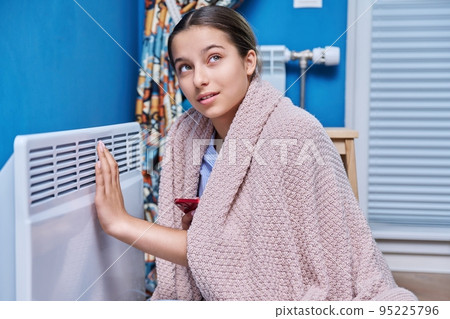 Young female sitting near an electric heater at home Young female sitting near an electric heater at home 95225796