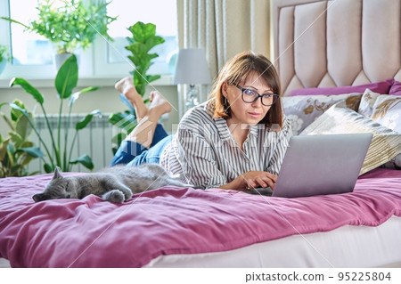 Woman together with cat lying on bed at home, female using laptop 95225804