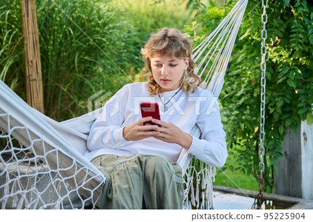 Teenage girl sitting in hammock using smartphone for leisure study communication 95225904