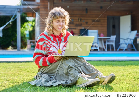 Teenage girl sitting on lawn in backyard, using laptop for studying leisure 95225906