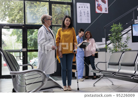 Senior physician helping injured patient to walk with stick, suffering from physical impairment and using cane. Doctor attending rehabilitation therapy to recover from leg injury at facility. 95226009