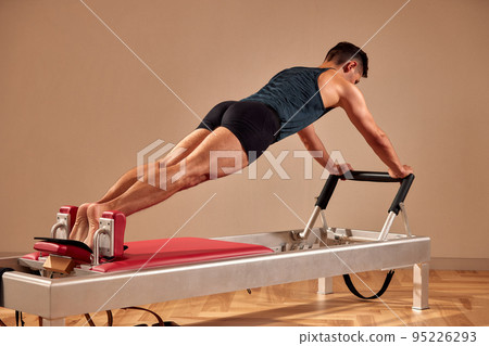 Side view of barefoot male athlete lying on pilates reformer and performing abs exercise during fitness workout. Pilates man concept. 95226293