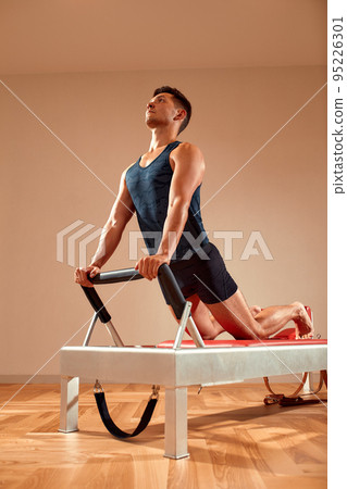 Fit man doing a lunge stretch yoga pilates exercise to strengthen and tone his muscles using a reformer in a gym Fit man doing a lunge stretch yoga pilates exercise to strengthen and tone his muscles using a reformer in a gym 95226301