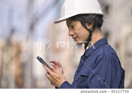 A man in work clothes looking at a smartphone in front of a construction site A man in work clothes looking at a smartphone in front of a construction site 95227750