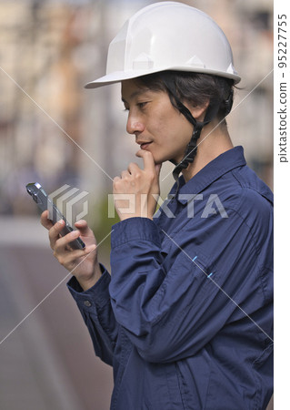 A man in work clothes looking at a smartphone in front of a construction site A man in work clothes looking at a smartphone in front of a construction site 95227755