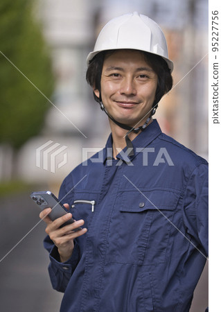 A man in work clothes looking at a smartphone in front of a construction site A man in work clothes looking at a smartphone in front of a construction site 95227756