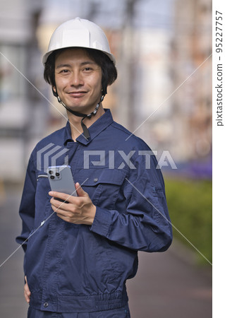 A man in work clothes looking at a smartphone in front of a construction site A man in work clothes looking at a smartphone in front of a construction site 95227757