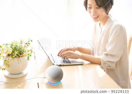 A young woman talking to an AI speaker while working on a PC 95229133
