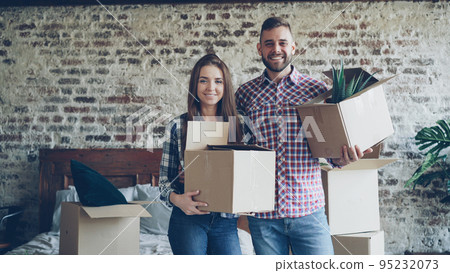 Portrait of happy young couple standing in bedroom in new house, holding carton boxes, smiling and looking at camera. Family, relationship and relocation concept. 95232073