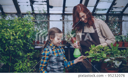 Curious little girl is helping her mom to spray green plants with water from spraybottle while working together in orchard. Childhood, people and growing flowers concept. 95232076