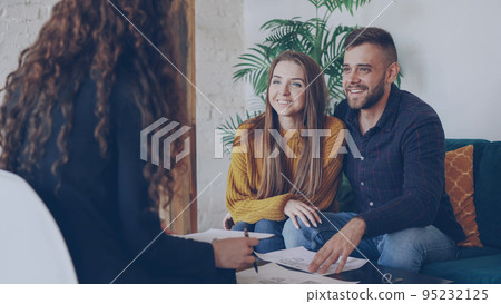Loving boyfriend and girlfriend looking at plan of house they are going to buy and talking to real estate broker sitting at coffee table on comfortable couch 95232125