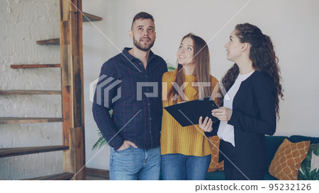 Cheerful house agent is talking to buyers adorable young couple discussing conditions of sale and showing papers. Smiling clients are excited about new apartment. 95232126
