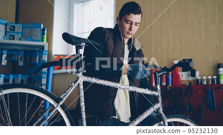 Professional serviceman is fixing bicycle handle-bar turning allen key to tighten joint and listening to music with earphones. Special tools, spares on shelves and equipment are visible. 95232130