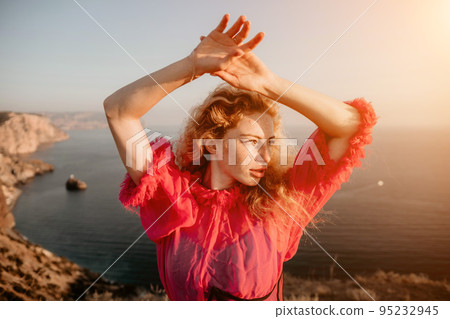 Close up portrait of curly redhead young caucasian woman with freckles looking at camera and smiling. Cute woman portrait in a pink long dress posing on a volcanic rock high above the sea at sunset Close up portrait of curly redhead young caucasian woman with freckles looking at camera and smiling. Cute woman portrait in a pink long dress posing on a volcanic rock high above the sea at sunset 95232945