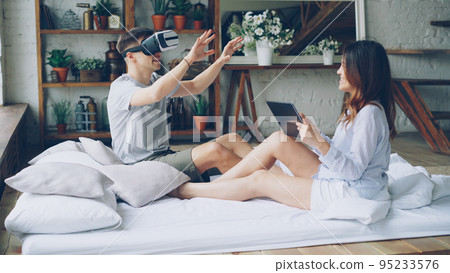 Young man is using virtual reality glasses while his attractive wife is touching tablet screen, looking at her husband and smiling. Modern technology and human relationship concept. 95233576