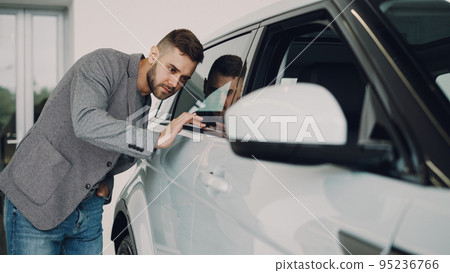 Stylish bearded guy is choosing new automobile walking around luxury car and touching it in car dealership. Rich people, transportation and business class vehicles concept. Stylish bearded guy is choosing new automobile walking around luxury car and touching it in car dealership. Rich people, transportation and business class vehicles concept. 95236766