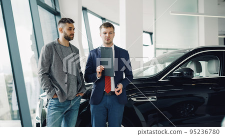 Handsome bearded guy is talking to car dealership manager in motor show discussing new automobile model, dealer is holding documents and pen. Purchasing vehicle and people concept. 95236780