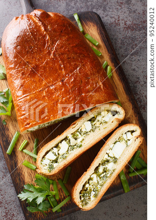 Baked yeast pie stuffed with boiled eggs, green onions, parsley, dill and spinach close-up on a wooden board. Vertical top view 95240012