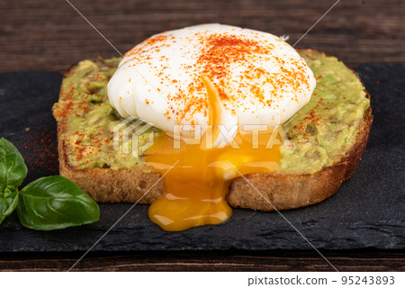Avocado toast with leaking poached egg yolk on a stone board for serving. 95243893