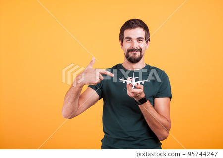Travel concept image of man portrait pointing to a toy airplane that he holds in his hands isolated on orange background in studio. Vacation, voyage and summer journey image Travel concept image of man portrait pointing to a toy airplane that he holds in his hands isolated on orange background in studio. Vacation, voyage and summer journey image 95244247