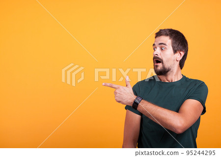 Expressive shocked man pointing at his right with his mouth open on orange background. Surprised guy pointing at the copyspace which is available for your advertising or promotions Expressive shocked man pointing at his right with his mouth open on orange background. Surprised guy pointing at the copyspace which is available for your advertising or promotions 95244295