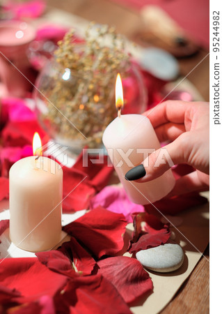 female hand holding burning candle over rose petals, stones and herbs layered on a table. aura readings and magical rituals, spiritualistic and channeling session. 95244982