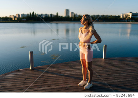 Sporty young woman exercising and stretching neck for warming up before running or working out near water on background of cityscape. Blonde sportswoman exercising in soft sunlight of summer morning. 95245694