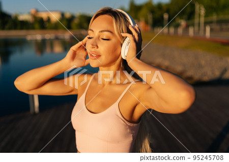 Close-up face of young blonde woman with closed eyes in workout sportswear putting on wireless headphones to start running in city park on summer morning. Female runner wearing wireless earphones. Close-up face of young blonde woman with closed eyes in workout sportswear putting on wireless headphones to start running in city park on summer morning. Female runner wearing wireless earphones. 95245708