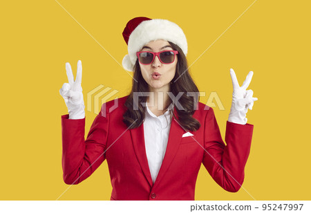 Surprised woman wearing in Santa style shows victory gesture with two hands on yellow background. 95247997