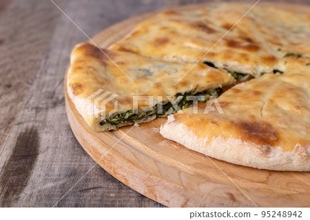 Pie with spinach, herbs and suluguni cheese on a wooden board. Whole pie with cut off piece. Traditional round flat pie with filling. Homemade pastries of Caucasian and Balkarian cuisine. Pie with spinach, herbs and suluguni cheese on a wooden board. Whole pie with cut off piece. Traditional round flat pie with filling. Homemade pastries of Caucasian and Balkarian cuisine. 95248942
