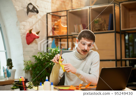 Young man sitting at home, holds a master class on origami and recording vlog at home studio. Hobbies and leisure activities. Remote training, online classes 95249430