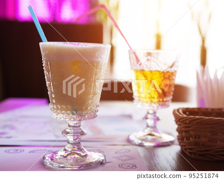 Two glasses with drinks in a restaurant, a coconut cocktail with a straw and apple juice against the backdrop of window light. Summer drinks. Two glasses with drinks in a restaurant, a coconut cocktail with a straw and apple juice against the backdrop of window light. Summer drinks. 95251874