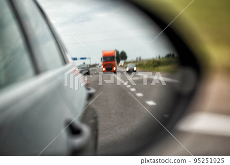 View from the side rear-view mirror on the traffic situation. Truck and cars with headlights on. Traffic on the road, road safety. Blurred, close-up View from the side rear-view mirror on the traffic situation. Truck and cars with headlights on. Traffic on the road, road safety. Blurred, close-up 95251925
