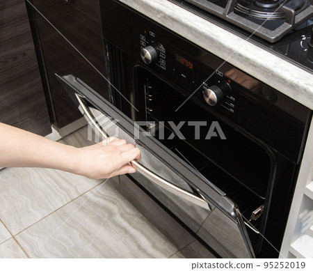 A woman's hand opens the door of a modern electric oven in the kitchen. Copy space for text A woman's hand opens the door of a modern electric oven in the kitchen. Copy space for text 95252019