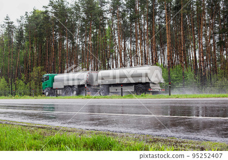 A truck with a tank semi-trailer transports liquid cargo on a slippery wet road in summer against the backdrop of a forest. Transportation of goods in bad weather. Sanctions A truck with a tank semi-trailer transports liquid cargo on a slippery wet road in summer against the backdrop of a forest. Transportation of goods in bad weather. Sanctions 95252407