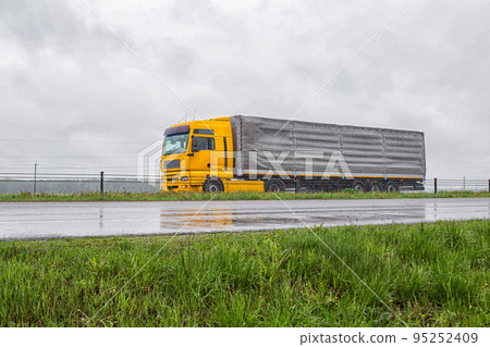 A yellow modern truck with a tilt semi-trailer carries cargo along the highway in cloudy rainy weather. Industry A yellow modern truck with a tilt semi-trailer carries cargo along the highway in cloudy rainy weather. Industry 95252409