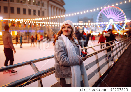 Smiling woman in winter style clothes with coffee near skating rink. Young woman enjoying winter holidays. Smiling woman in winter style clothes with coffee near skating rink. Young woman enjoying winter holidays. 95257203