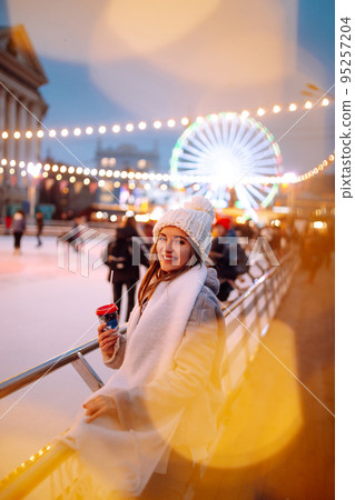 Smiling woman in winter style clothes with coffee near skating rink. Young woman enjoying winter holidays. Smiling woman in winter style clothes with coffee near skating rink. Young woman enjoying winter holidays. 95257204